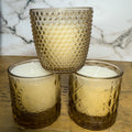 Three textured glass candle holders with candles on a wooden surface.