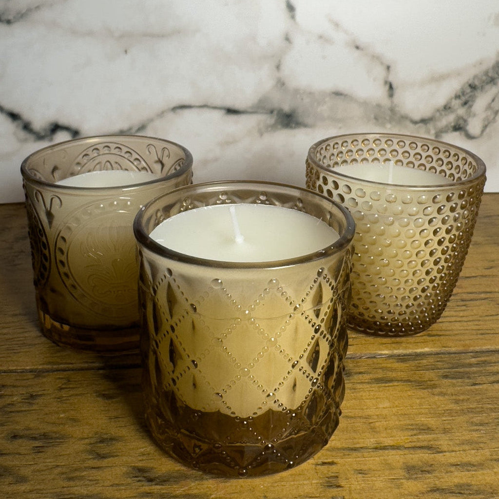 Three textured glass candle holders with candles on a wooden surface against a marble wall.
