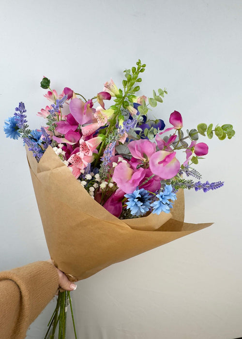 A bouquet of colourful cottage garden faux flowers wrapped in brown paper held by a person against a plain background