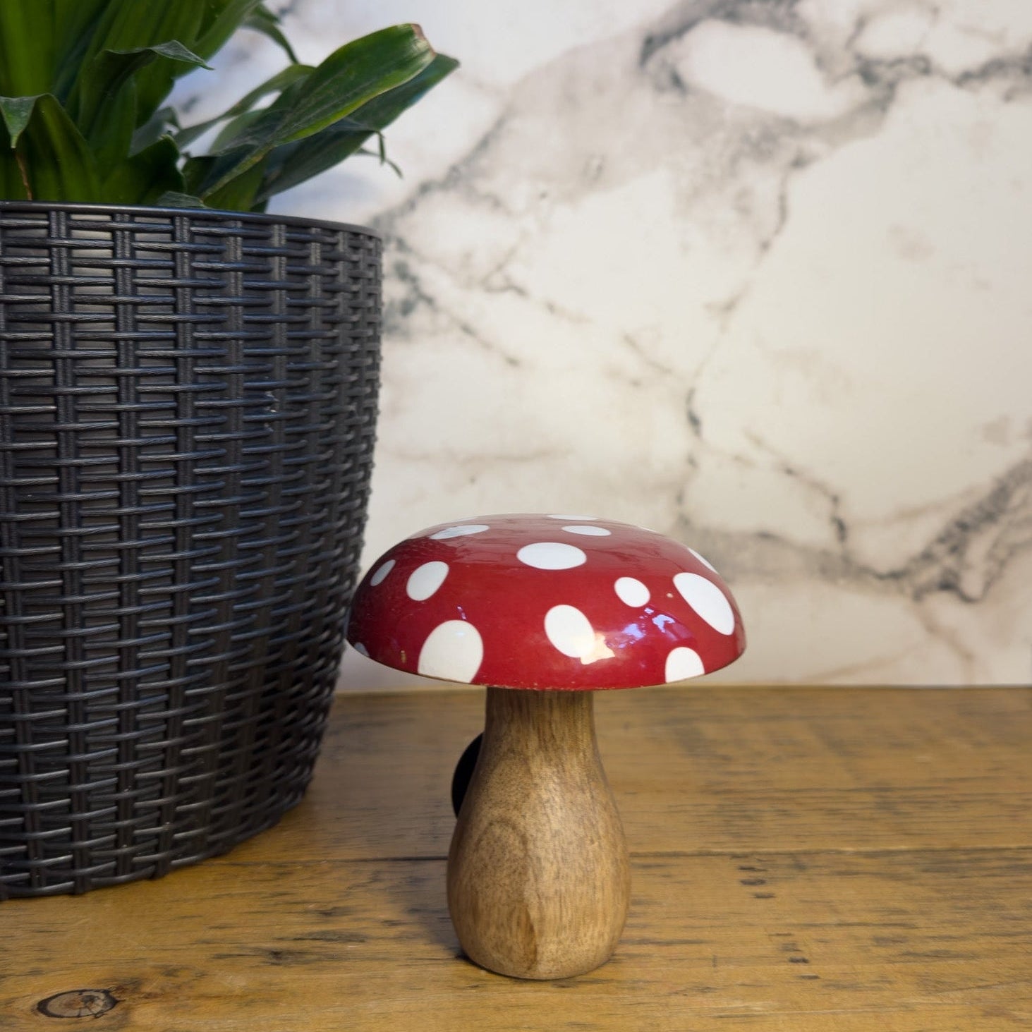 Wooden mushroom with red cap and white spots on a wooden surface next to a plant and marble wall.