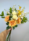 A bouquet of creamy yellow, white and orange realistic faux flowers held by a person against a light blue background. Includes Dahlias, roses and dancing orchids and foliage