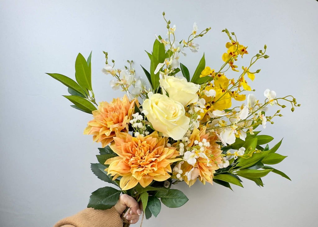 A bouquet of creamy yellow, white and orange realistic faux flowers held by a person against a light blue background. Includes Dahlias, roses and dancing orchids and foliage